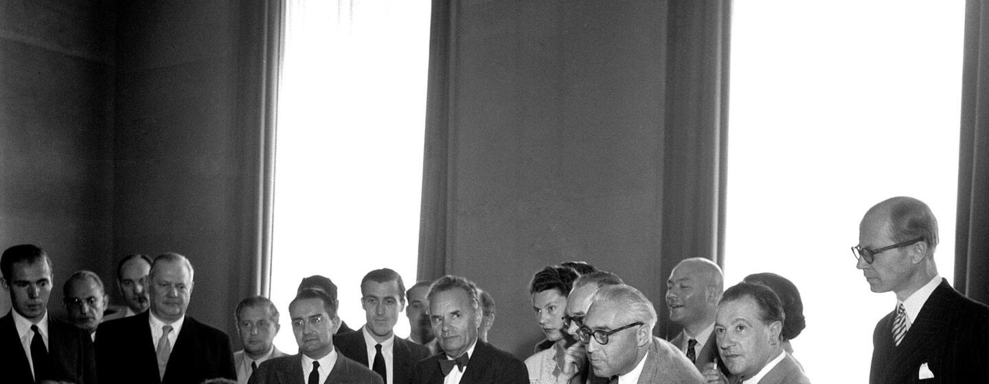 Autoridades no Palácio das Nações, em Genebra, assinam a Convenção Relativa ao Estatuto dos Refugiados em 1951. Sentados à mesa estão, da esquerda para a direita, Margaret Kitchen, John Humphrey, Knud Larsen e o Dr. G.J. Van Heuven Goedhart, com outros delegados e observadores em pé atrás deles.