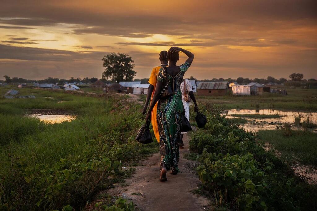 Au coucher du soleil, un groupe de femmes et d'enfants marchent le long d'un chemin dans un village rural, avec des champs inondés et des abris rudimentaires en arrière-plan.