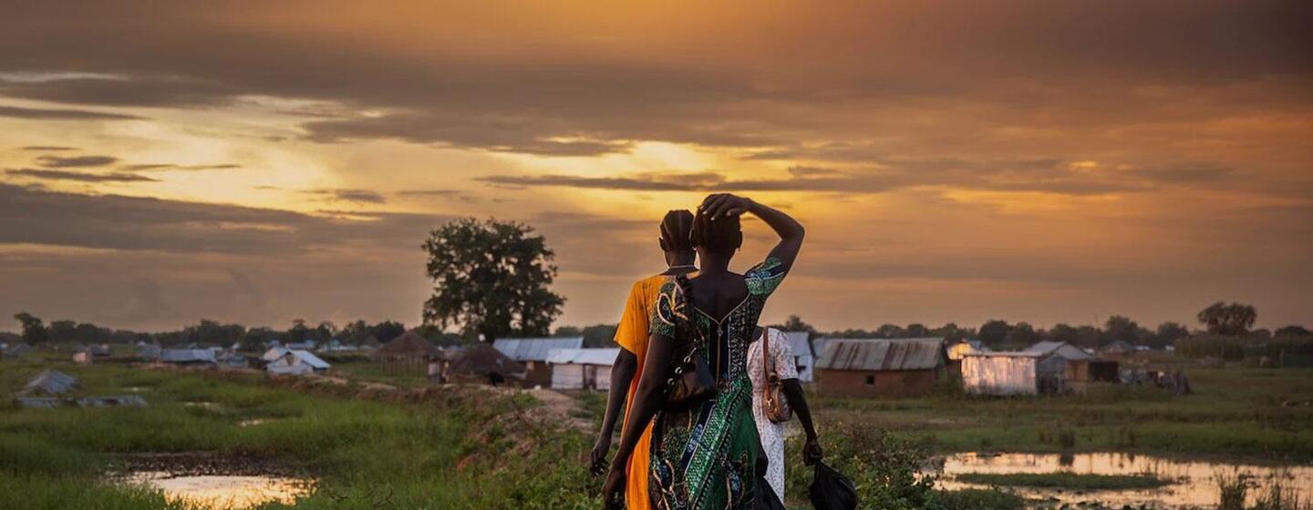 A group of women and children walk along a path in a rural village at sunset, with flooded fields and simple shelters in the background.