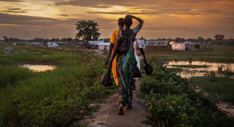 Au coucher du soleil, un groupe de femmes et d'enfants marchent le long d'un chemin dans un village rural, avec des champs inondés et des abris rudimentaires en arrière-plan.