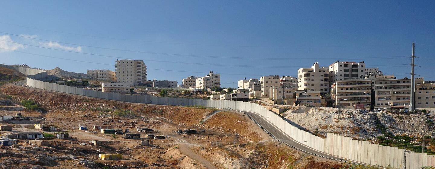 Une clôture de sécurité israélienne longe un flanc de colline, séparant un chantier de construction au premier plan d'une zone résidentielle aux bâtiments blancs perchés sur une colline à l'arrière-plan, sous un ciel bleu dégagé.