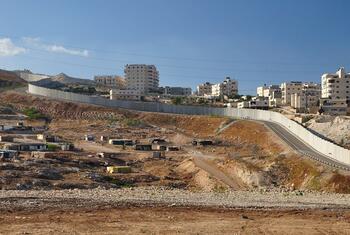 Une clôture de sécurité israélienne longe un flanc de colline, séparant un chantier de construction au premier plan d'une zone résidentielle aux bâtiments blancs perchés sur une colline à l'arrière-plan, sous un ciel bleu dégagé.