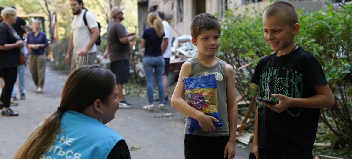 An aid worker in a blue vest talks with two young boys in a damaged residential area in Kyiv, Ukraine, as part of UNOCHA's relief efforts.