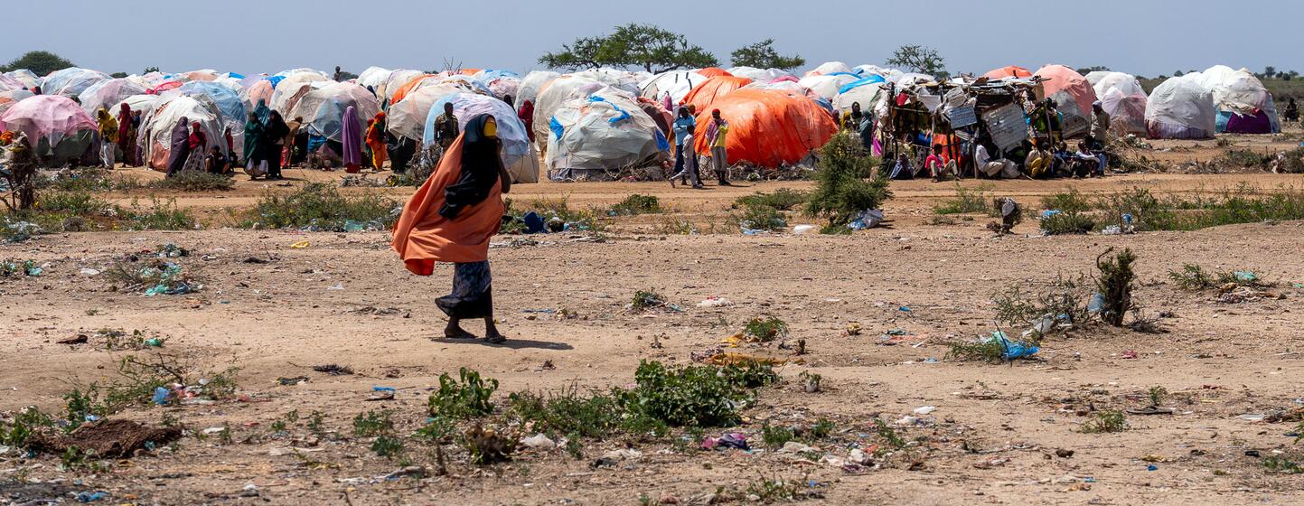 Una mujer con un pañuelo naranja brillante camina por un paisaje árido y polvoriento en un campamento de desplazados internos en Burhakaba, Somalia. Al fondo, numerosos refugios improvisados ​​construidos con grandes lonas de colores se agrupan bajo un cielo brillante y parcialmente nublado.