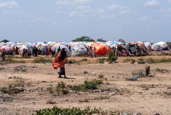 A woman in a vibrant orange headscarf walks through a dusty, arid landscape in an IDP camp in Burhakaba, Somalia. In the background, numerous makeshift shelters constructed from large, colorful tarps are clustered together under a bright, partly cloudy sky.