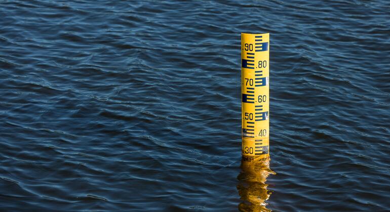 Un indicador de nivel de agua amarillo con marcas negras se alza en medio de una masa de agua azul ondulante.