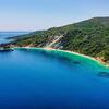 Vista aérea de la playa de Gidaki en Ítaca, Grecia, que muestra una bahía de color turquesa con una playa de arena y yates de lujo, rodeada de colinas verdes.
