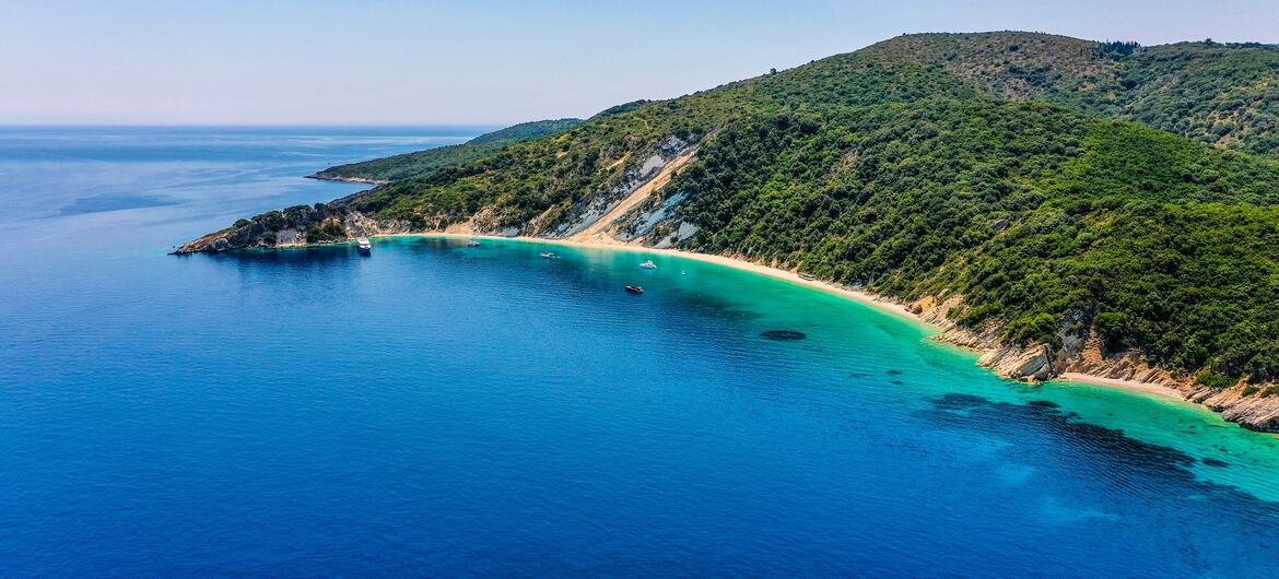 Aerial view of Gidaki Beach in Ithaca, Greece, showing a turquoise bay with a sandy beach and luxury boats, surrounded by green hills.