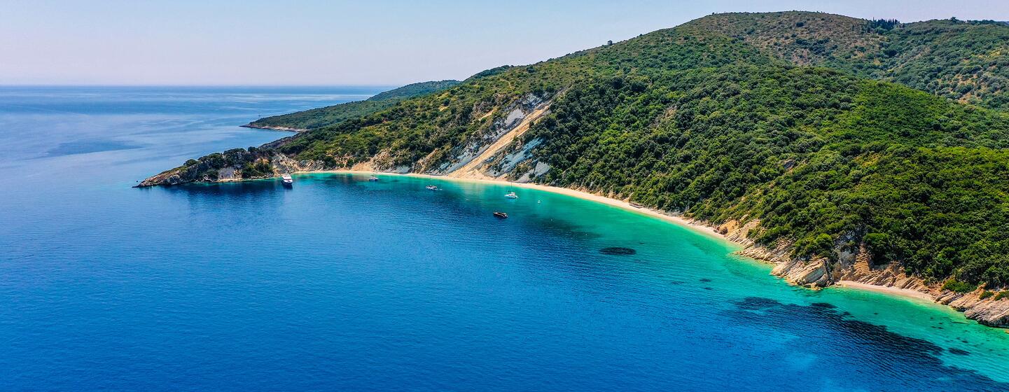 Vista aérea de la playa de Gidaki en Ítaca, Grecia, que muestra una bahía de color turquesa con una playa de arena y yates de lujo, rodeada de colinas verdes.