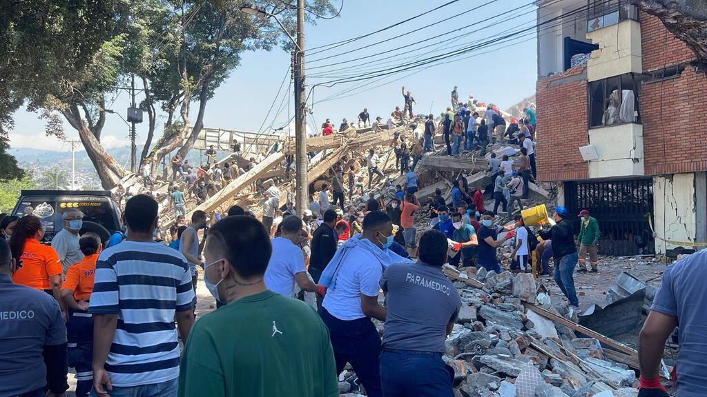 Watu wamesimama kati ya vifusi na jengo lililoanguka huku waokoaji wakipekua eneo hilo, huku kuta za matofali zilizoharibika na mihimili iliyoanguka ikiwa nyuma.