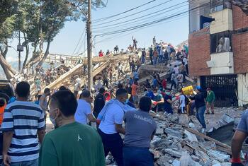 People stand among rubble and a collapsed building as rescuers search the site, with damaged brick walls and fallen beams in the background.