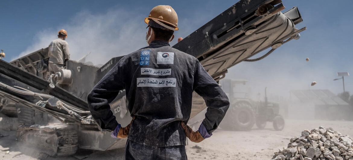 A UNDP worker in a hard hat and work clothes stands with hands on hips watching a machine clear rubble during Syria's post-conflict recovery efforts.