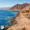 Vue aérienne d'une côte ensoleillée de la mer Rouge à Dahab, en Égypte, avec une eau bleue, des rivages rocheux, des montagnes désertiques et des personnes faisant de la plongée en apnée près d'un petit bateau.