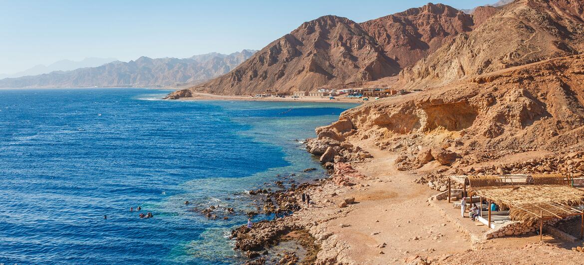 Vue aérienne d'une côte ensoleillée de la mer Rouge à Dahab, en Égypte, avec une eau bleue, des rivages rocheux, des montagnes désertiques et des personnes faisant de la plongée en apnée près d'un petit bateau.