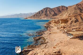 Vista aérea de una soleada costa del Mar Rojo en Dahab, Egipto, con aguas azules, costas rocosas, montañas desérticas y gente practicando snorkel cerca de una pequeña embarcación.
