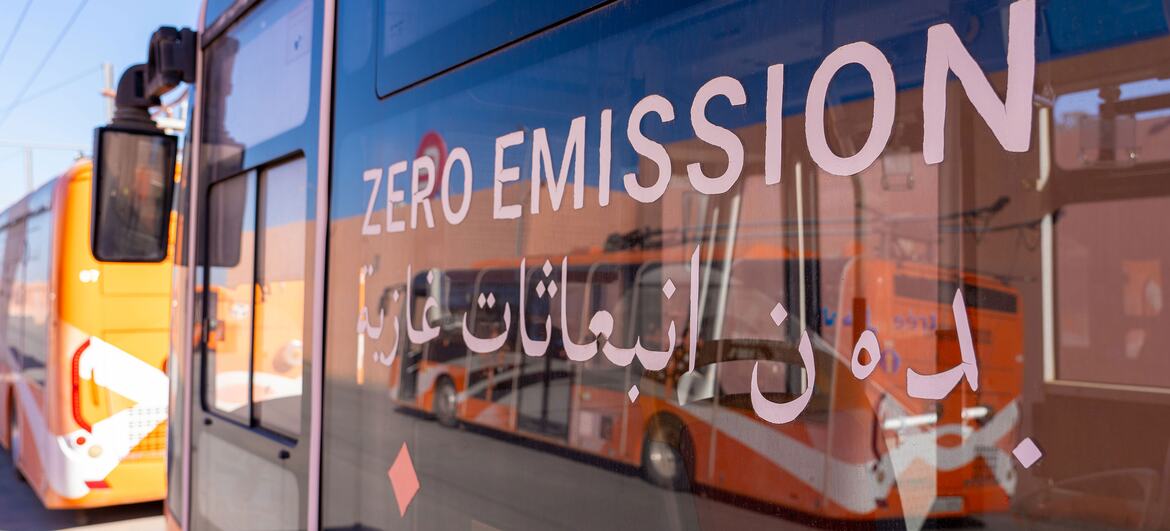 Primer plano de la ventana de un autobús eléctrico que muestra la inscripción "CERO EMISIONES" en inglés y árabe, con otro autobús naranja visible al fondo.