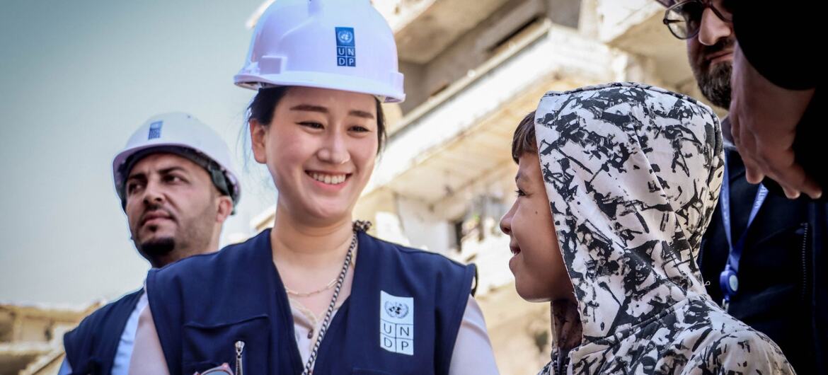 A smiling UNDP worker in a hard hat and vest speaks with a young boy in a camouflage hoodie during Syria's reconstruction efforts.