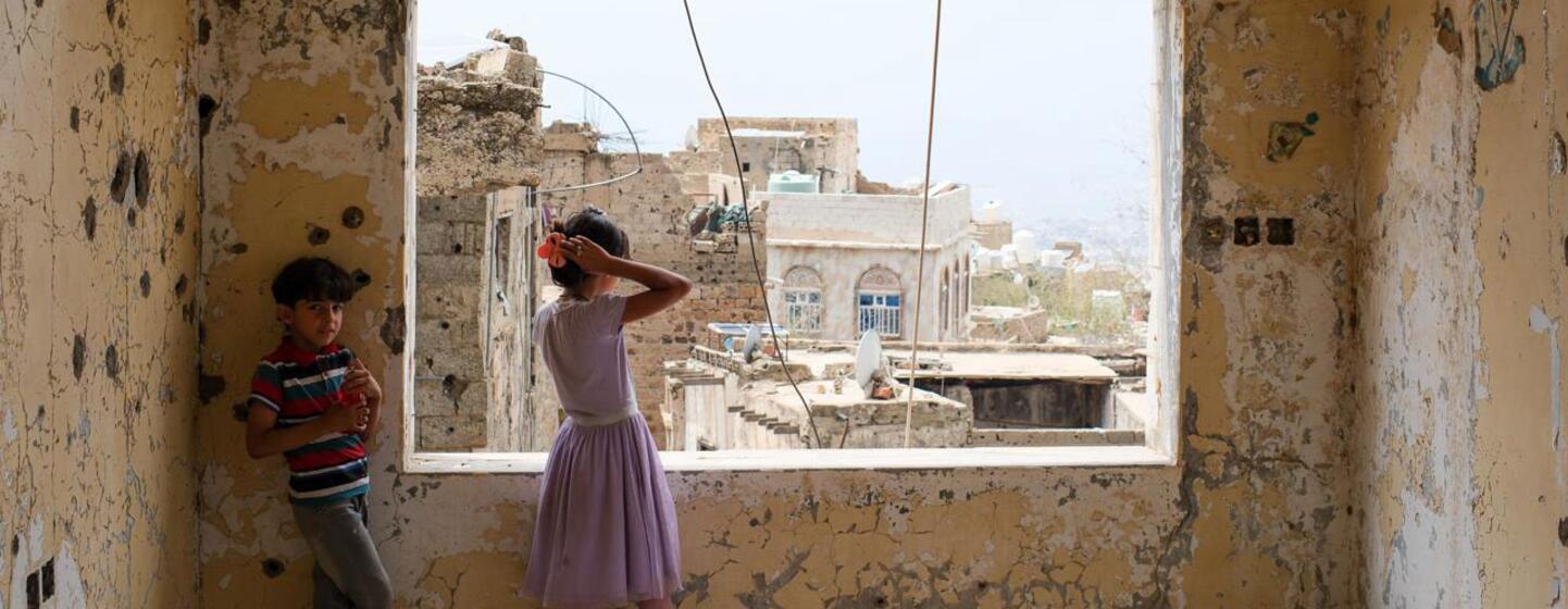 Deux enfants se tiennent debout dans une pièce endommagée, aux murs criblés de balles, regardant par une fenêtre cintrée la ville de Taïz, au Yémen.
