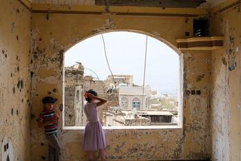 Two children stand in a damaged room with bullet holes on the walls, looking out of an arched window over the city of Taiz, Yemen.