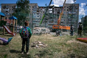 A young boy stands in a playground looking at a heavily damaged apartment building with cranes clearing rubble after an attack in Kyiv.