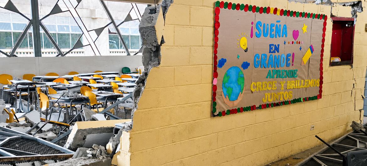 Uma sala de aula danificada na escola industrial La Carrasquilla, em Quibdó, Colômbia, mostrando uma parede de tijolos amarelos desabada e destroços espalhados entre as carteiras tombadas.
