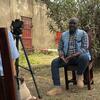 Emmanuel Madragule est assis à l'extérieur pendant qu'il est interviewé par une journaliste munie d'une caméra à Bunia, dans l'Ituri, en République démocratique du Congo.