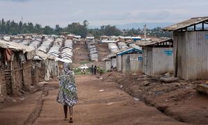 A woman in a colorful patterned dress walks down a dirt path through rows of makeshift shelters in a refugee camp in Ituri Province, DRC.