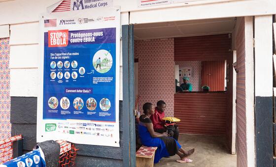 A large blue 'STOP EBOLA' informational poster hangs on a wall next to two young people sitting on a bench inside the Kigonze Ebola Transit Centre in DR Congo.