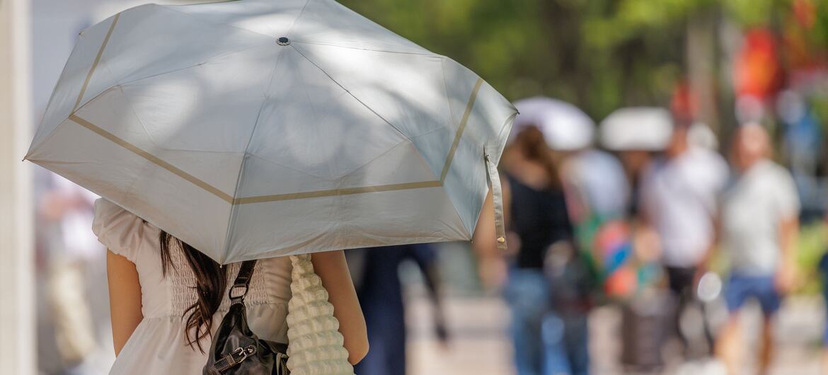 Japan’s ‘2050 Weather Forecast’ highlights extreme heat in the future 2 A woman in a white dress holds a large white parasol while walking on a sunny city street with other people in the background.