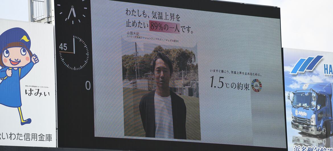 A large screen at a football stadium displays a video message from Yamada Taisuke about stopping temperature rise and the 1.5-degree promise during a match. Six J.LEAGUE Climate Action Ambassadors have recorded video messages for the initiative, including Hiroki Yamada, a long-time star of Júbilo Iwata. In his message, Mr. Yamada expresses concern that children may increasingly be unable to play outdoors during summer and emphasizes the role individuals can play in addressing climate change. His message was shown on the giant screen at Júbilo Iwata's opening home match of the 2026/27 season on 8 August. The match kicked off at 7 pm as part of league-wide efforts to reduce heat-related risks.