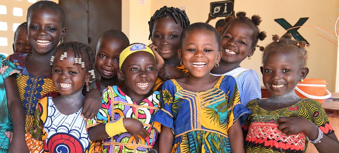 A group of smiling African preschool children in colorful clothing pose together inside a classroom.