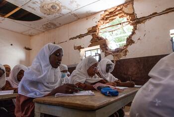 Female students in hijabs attend a lesson at Amina Bint Wahb Primary School for Girls in Khartoum, with visible damage to the walls and ceiling.