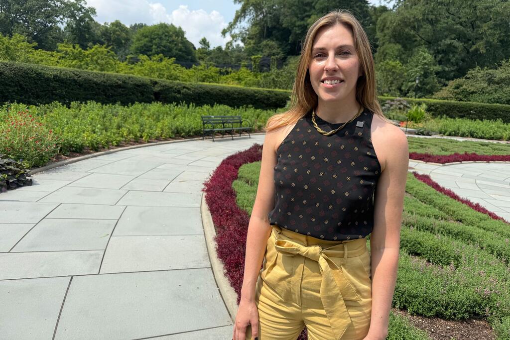 Una mujer sonriente de cabello rubio se encuentra de pie en un sendero pavimentado en un jardín paisajístico dentro de Central Park.