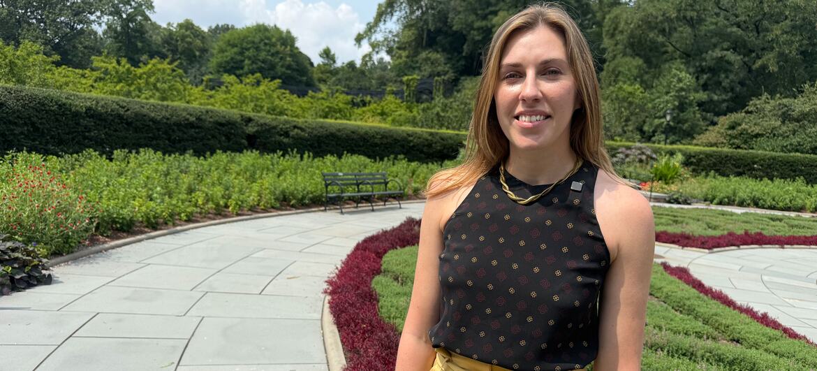 A smiling woman with blonde hair stands on a paved path in a landscaped garden within Central Park.