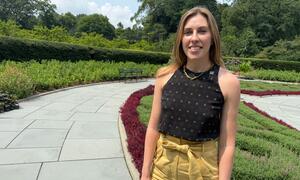 Une femme souriante aux cheveux blonds se tient sur un chemin pavé dans un jardin paysager de Central Park.