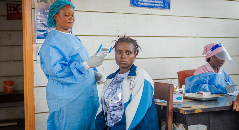 Uma parteira apoiada pelo UNFPA, vestindo equipamento de proteção azul, verifica a temperatura de uma mulher em um centro de saúde no campo de deslocados de Kigonze.