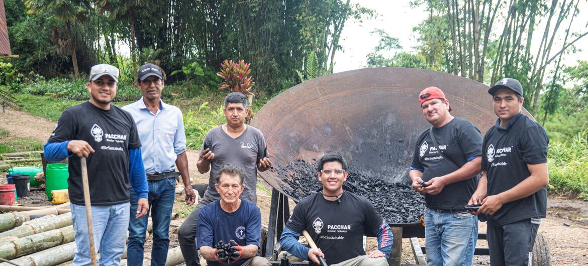 A group of men in Pacchar branded t-shirts pose with freshly made biochar and a large metal kiln in an outdoor, forested setting.