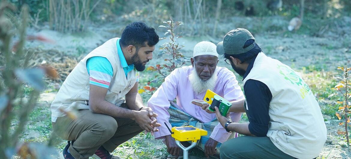 Two researchers in vests show a handheld device to an elderly farmer while examining a small yellow and green instrument on the ground during a land restoration project.