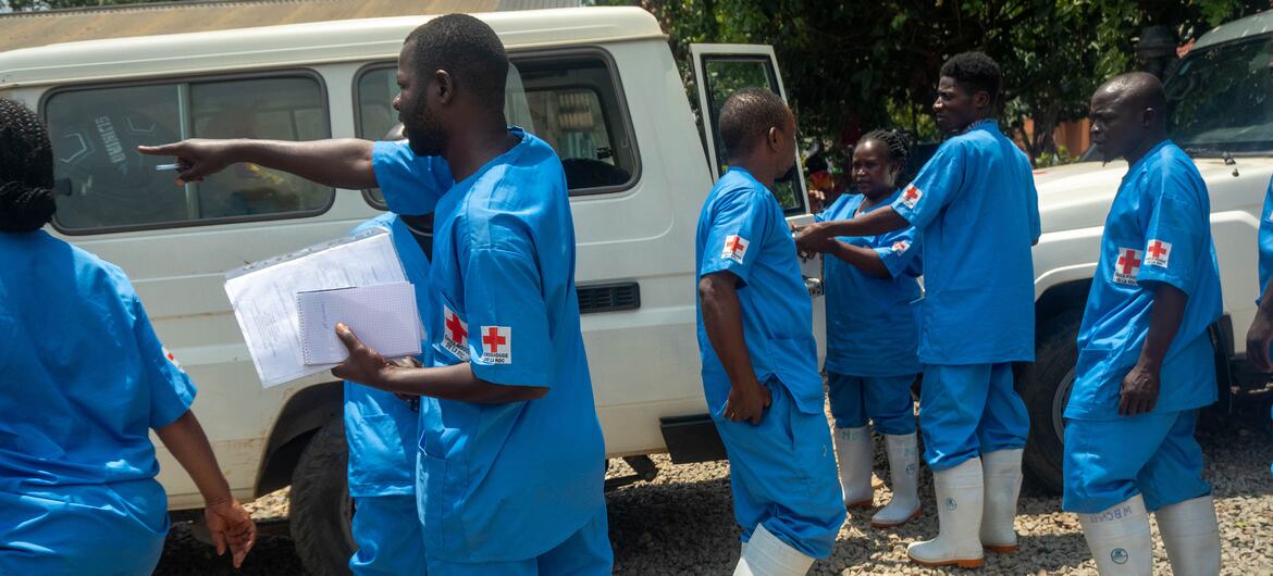 A team of DRC Red Cross volunteers in blue uniforms and white boots stands near a white van in Bunia, Ituri, preparing for an Ebola response mission.