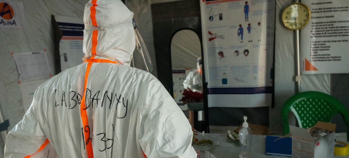 Medical worker in white PPE with orange stripes and 'LAB DANNY' written on the back stands at a table inside an Ebola treatment centre tent, with ALIMA posters, a wall clock, and medical supplies visible.