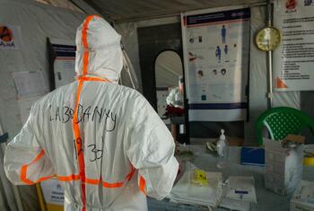 Medical worker in white PPE with orange stripes and 'LAB DANNY' written on the back stands at a table inside an Ebola treatment centre tent, with ALIMA posters, a wall clock, and medical supplies visible.