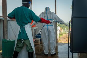 Un agent médical en blouse verte et visière pulvérise du désinfectant sur le dos d'un collègue portant une combinaison EPI blanche intégrale marquée « DÉCONTAMINE » dans un centre de traitement Ebola à Rwampara, Ituri, RDC.