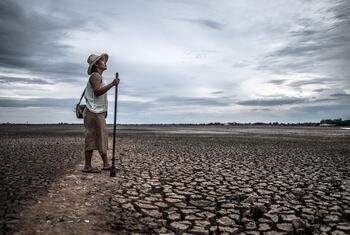 A woman wearing a straw hat and carrying a shoulder bag stands holding a long pole on a vast field of cracked, dried-up earth, with an old fishing boat resting on the parched ground behind her under a grey, overcast sky.