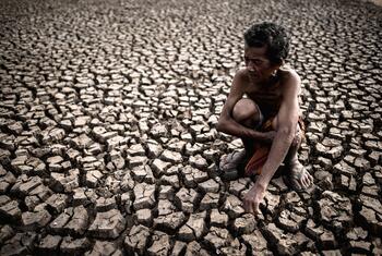 An elderly shirtless man crouches on a vast field of deeply cracked, dry earth.