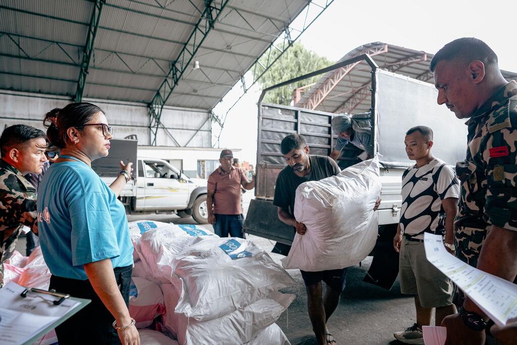 Un membre du personnel de UNICEF, vêtu d'une chemise bleue, coordonne ses actions avec des soldats et des ouvriers qui chargent des sacs blancs de matériel de secours sur un camion dans une zone de transit aéroportuaire.