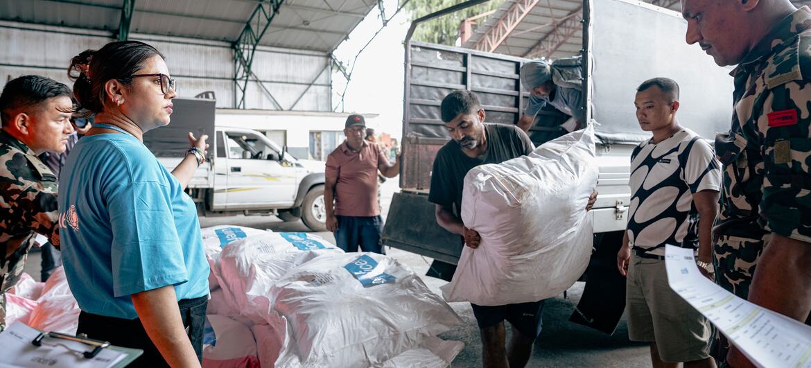A UNICEF staff member in a blue shirt coordinates with soldiers and workers loading white relief supply bags onto a truck at an airport staging area.