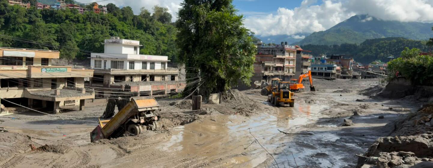 Un village recouvert de boue dans le district de Nuwakot, au Népal, avec des bâtiments endommagés et des engins de secours en action après les crues soudaines du 26 août 2026.