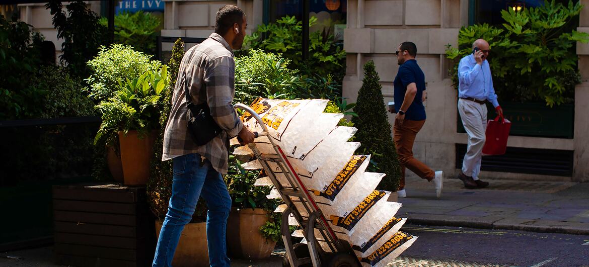 A delivery worker pushes a wheelbarrow loaded with stacked newspapers along a sunlit city sidewalk in the United Kingdom. Two men walk past in the background near potted plants and a stone building facade.