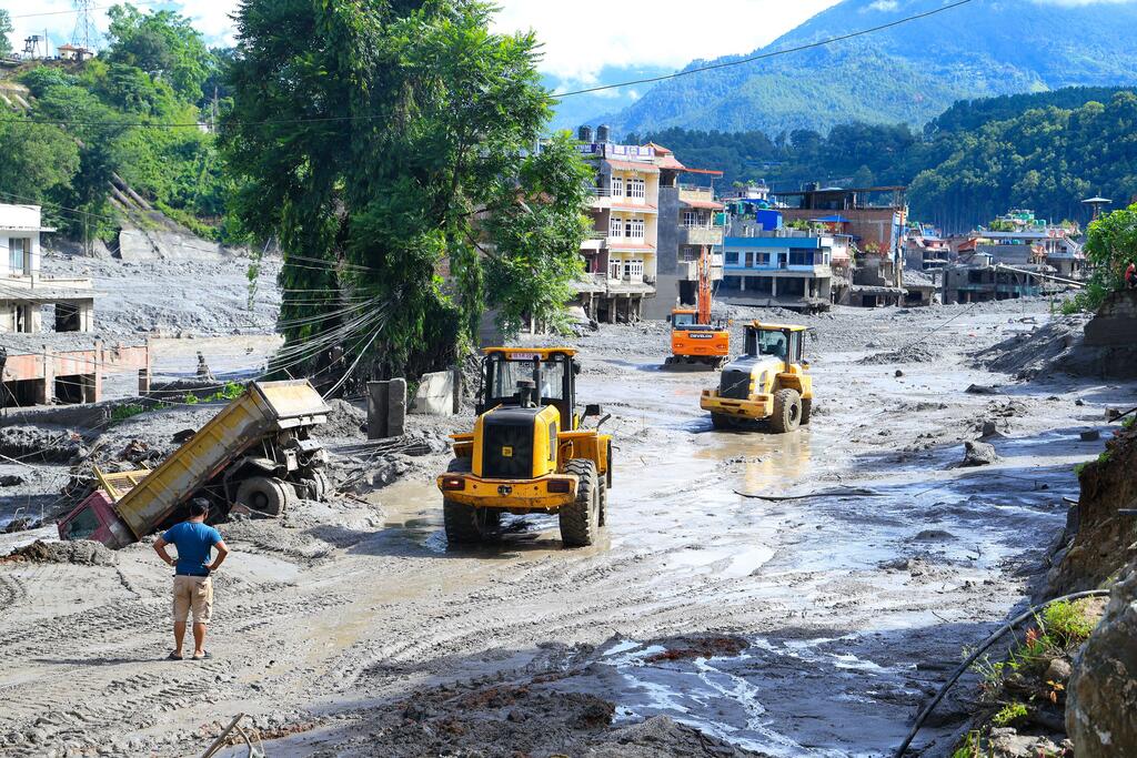 Des engins de chantier lourds, notamment des chargeuses et un camion-benne, sont à l'œuvre dans un village recouvert de boue à Nuwakot, au Népal, avec des bâtiments endommagés et des montagnes en arrière-plan après des crues soudaines.