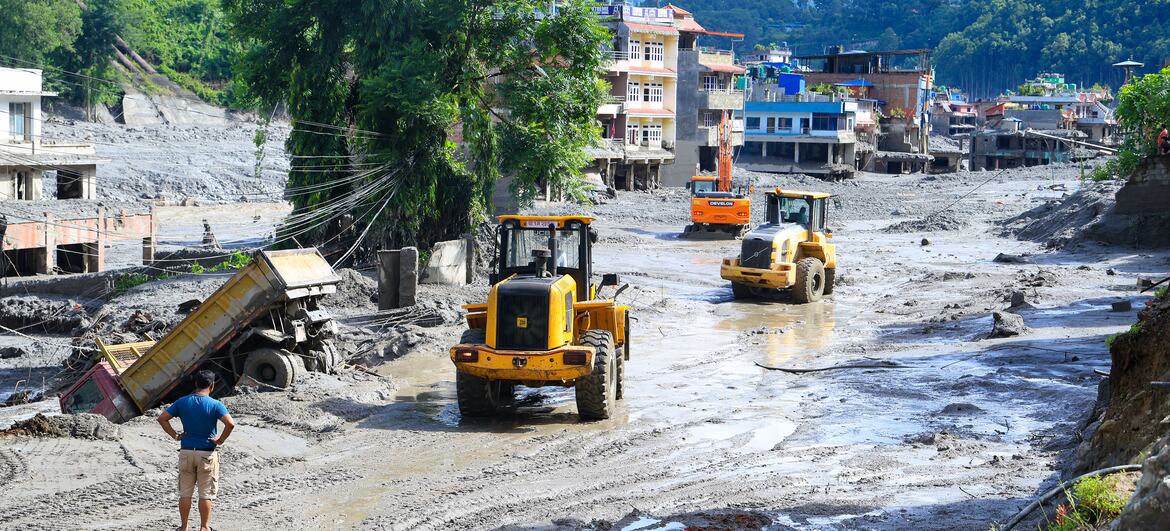 Equipamentos pesados ​​de construção, incluindo carregadeiras e um caminhão basculante, operam em um assentamento coberto de lama em Nuwakot, Nepal, com prédios danificados e montanhas ao fundo após enchentes repentinas.